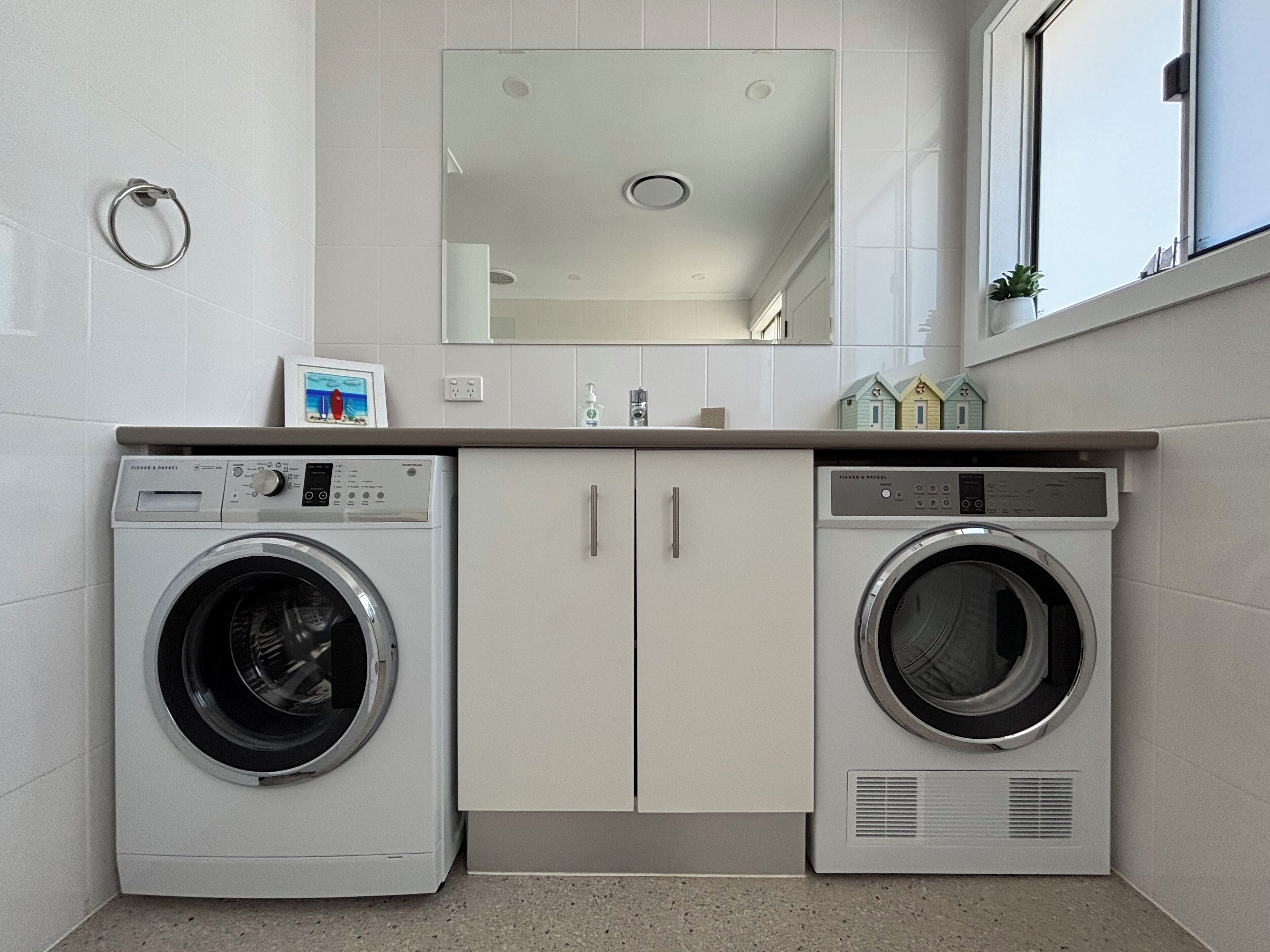 A laundry room with two front-loading washing machines, a mirror above the countertop, and a window with a plant.