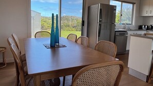 Un coin repas avec une table en bois et des chaises en osier, un réfrigérateur en acier inoxydable et une vue sur la verdure grâce à de grandes fenêtres.