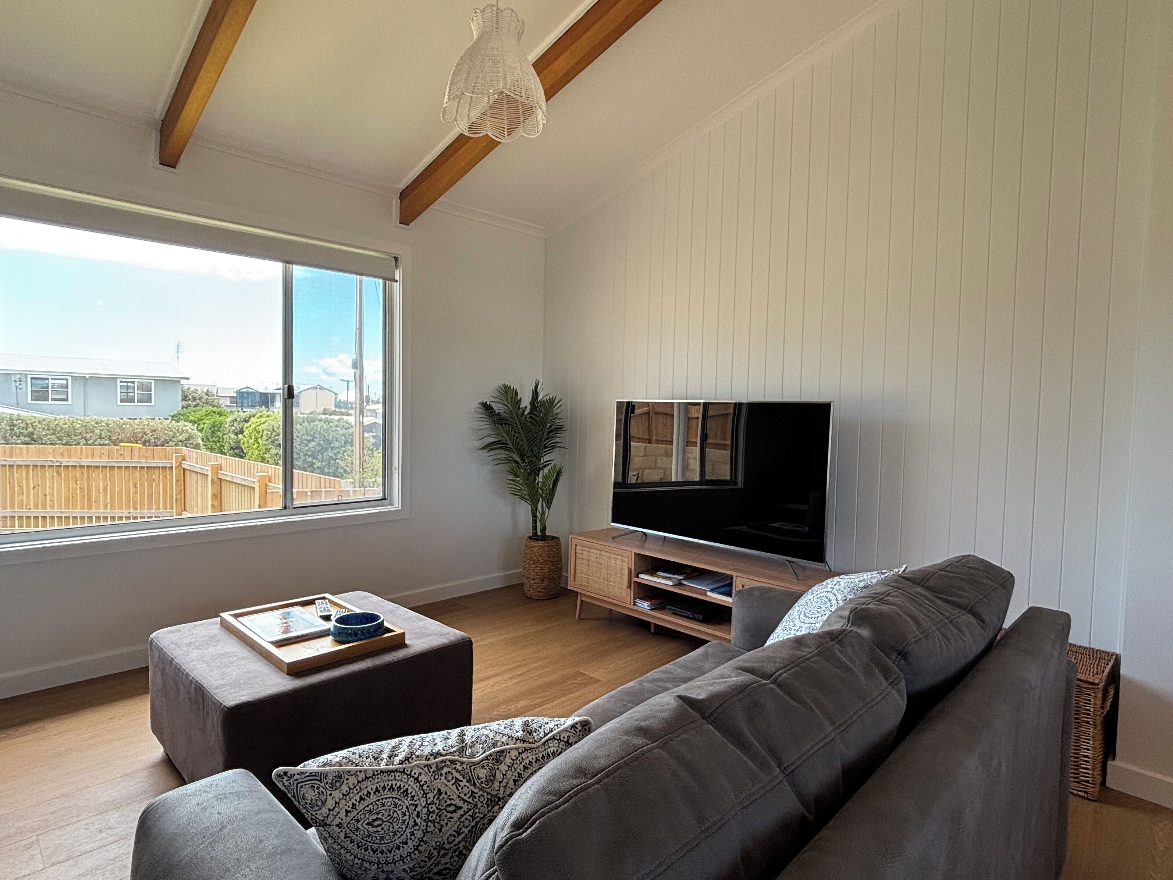 A modern living room with a large window, a flat-screen TV, a grey sofa, and a wooden ceiling.