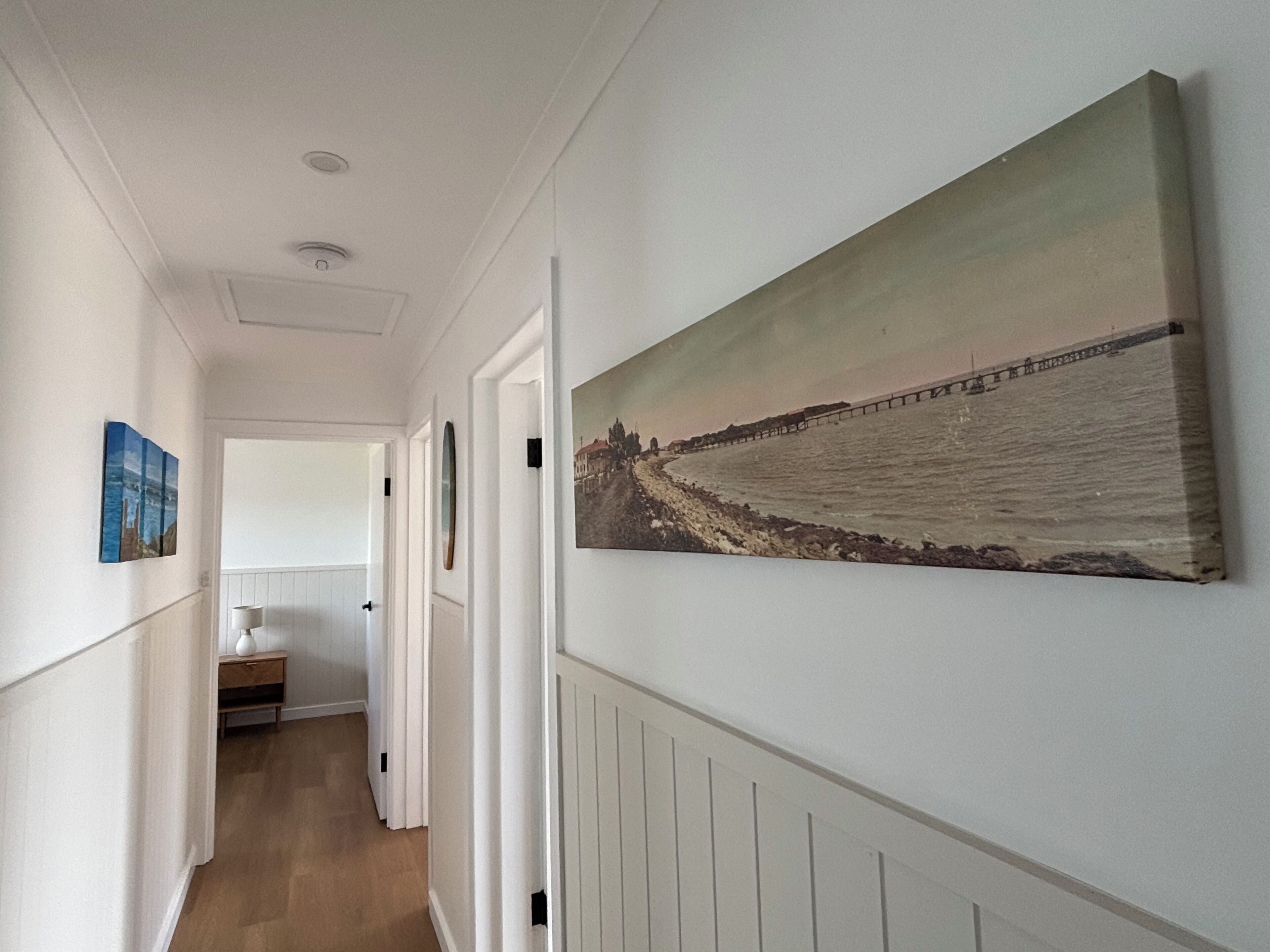 A hallway with a large painting of a pier and water, white walls, and a wooden floor.