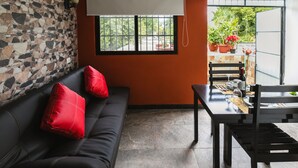 Un espace de vie moderne avec un canapé en cuir noir, des coussins rouges, une table à manger avec des chaises et une vue sur l’extérieur par une fenêtre.