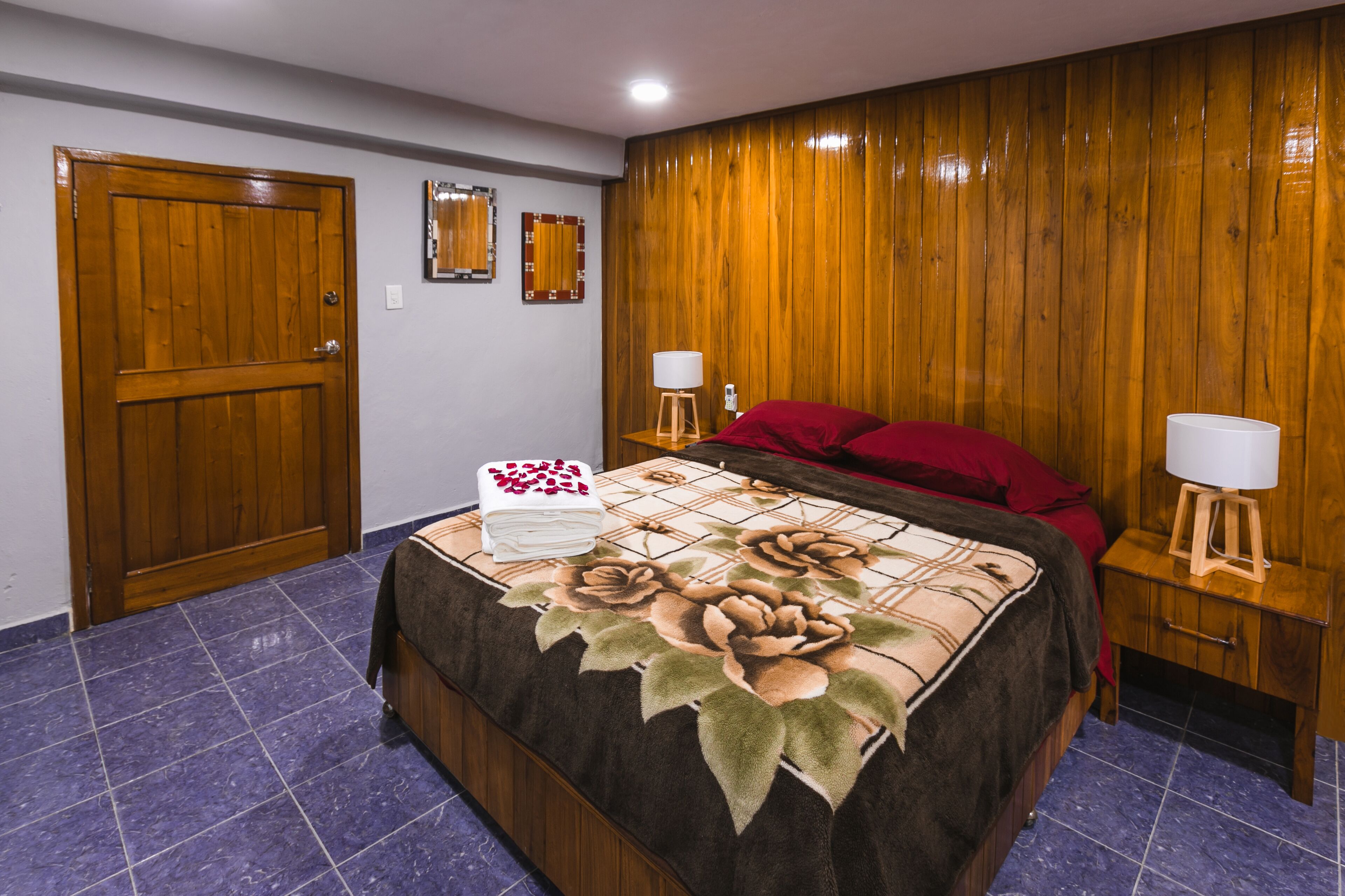 Une chambre avec une porte en bois, un lit recouvert d’une couvre-lit à motifs floraux, deux tables de chevet avec des lampes et un miroir accroché au mur.