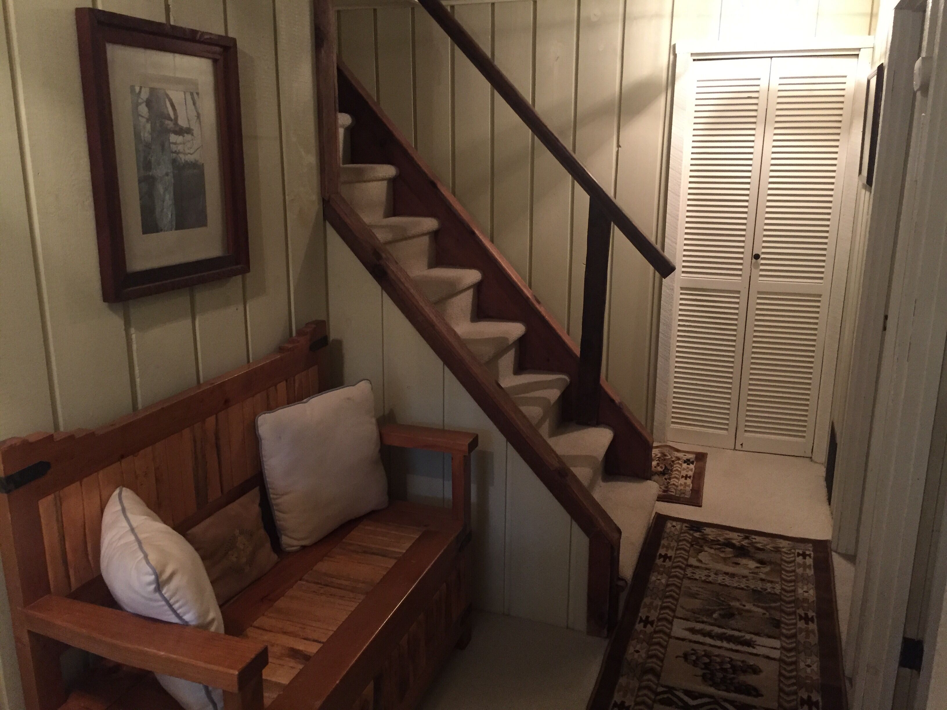 Eine Holztreppe mit Teppich und einer Sitzbank mit Kissen, sowie eine Tür mit verstellbaren Holzlamellen.