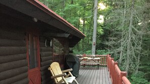 Una terraza de madera con una mecedora y una parrilla cubierta, rodeada por una valla de madera y un bosque denso.