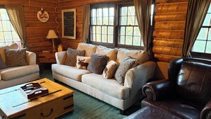 Interior de una cabaña de madera con un sofá, un sillón de cuero, un cofre de madera y una ventana con cortinas.