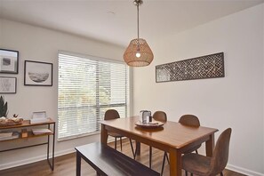 En matplats med ett träbord, fyra stolar, en bänk, en hängande, vävd lampa, ett fönster med persienner och en dekorativ väggdekoration.