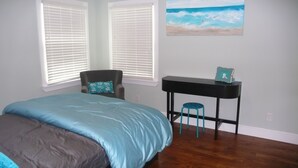 Een slaapkamer met een bed, een stoel, een bureau met krukje en een schilderij van een strandgezicht.