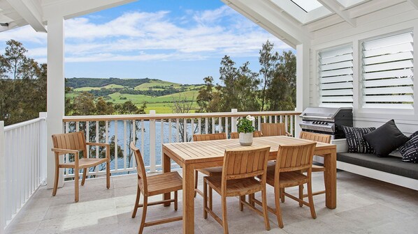 A covered outdoor patio with a wooden table and chairs, a grill, and a view of a green landscape. - Saltwater - walk to the beach (Werri Beach)