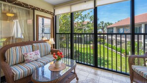 A balcony with a wicker sofa, a glass table with a vase of flowers, and a view of a green garden and buildings. - Sprinkle of Irish charm, beautifully updated, Siesta Dunes 8-205 (Siesta Key)