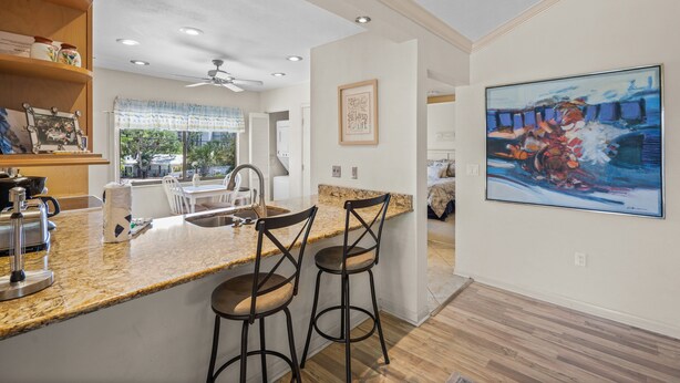A modern kitchen with granite countertops, two bar stools, a ceiling fan, and a large painting on the wall. - Sprinkle of Irish charm, beautifully updated, Siesta Dunes 8-205 (Siesta Key)