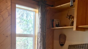 A cozy room with a wooden interior, a bed with a patterned blanket, a small table with a lamp, a shelf with books and decorative items, and a window with curtains. - the Stone Hut (Talkeetna)
