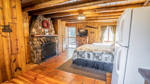 Interior acogedor de una cabaña con chimenea de piedra, televisor, cama y refrigerador.