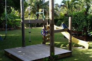Children can play on the slide and swing set amidst lush greenery and palm trees.