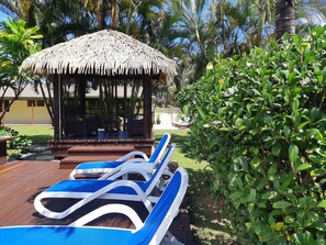 Un pabellón con techo de paja y asientos, rodeado de abundante vegetación y una terraza de madera con sillones azules.