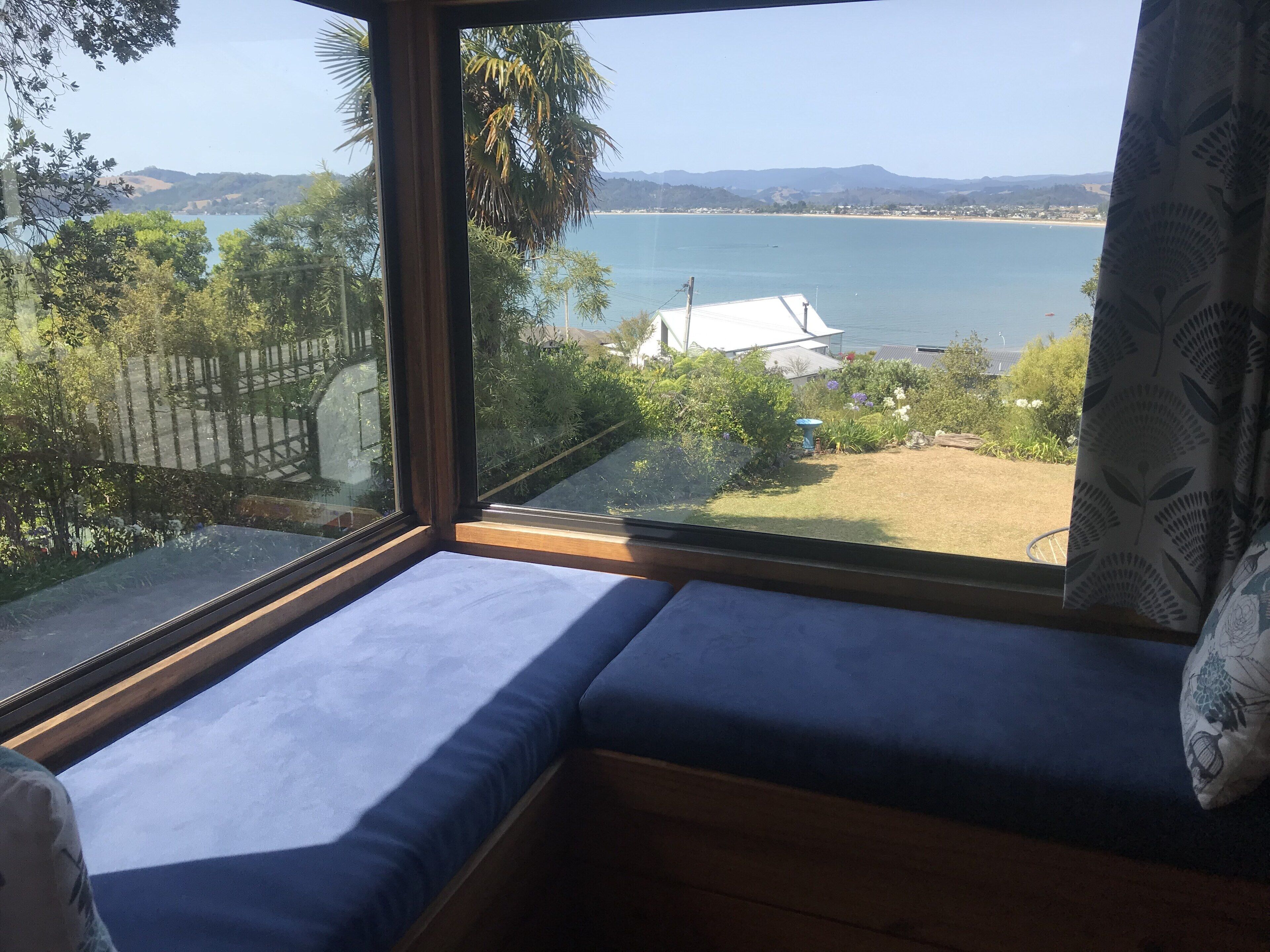 A room with a large window overlooking a coastal landscape, a blue cushioned bench, and a view of the sea and hills.