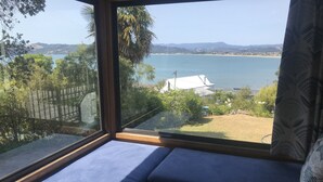A room with a large window overlooking a coastal landscape, a blue cushioned bench, and a view of the sea and hills.