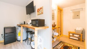 Una cocina compacta con refrigerador, microondas y lavaplatos. Hay una mesa de madera y taburetes de bar. Un televisor está montado en la pared.