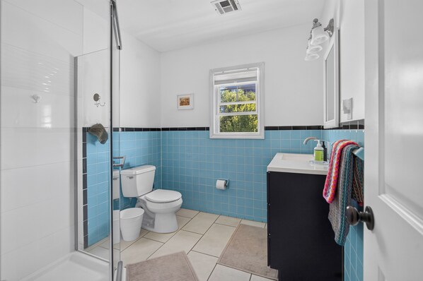 A bathroom with a glass shower enclosure, a toilet, a sink with a towel, and a window with a view of greenery.