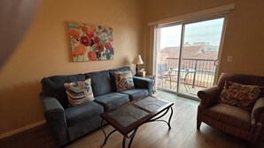 Uma sala de estar com sofá, poltrona e mesa de centro. Há um quadro na parede e uma janela com vista para edifícios.