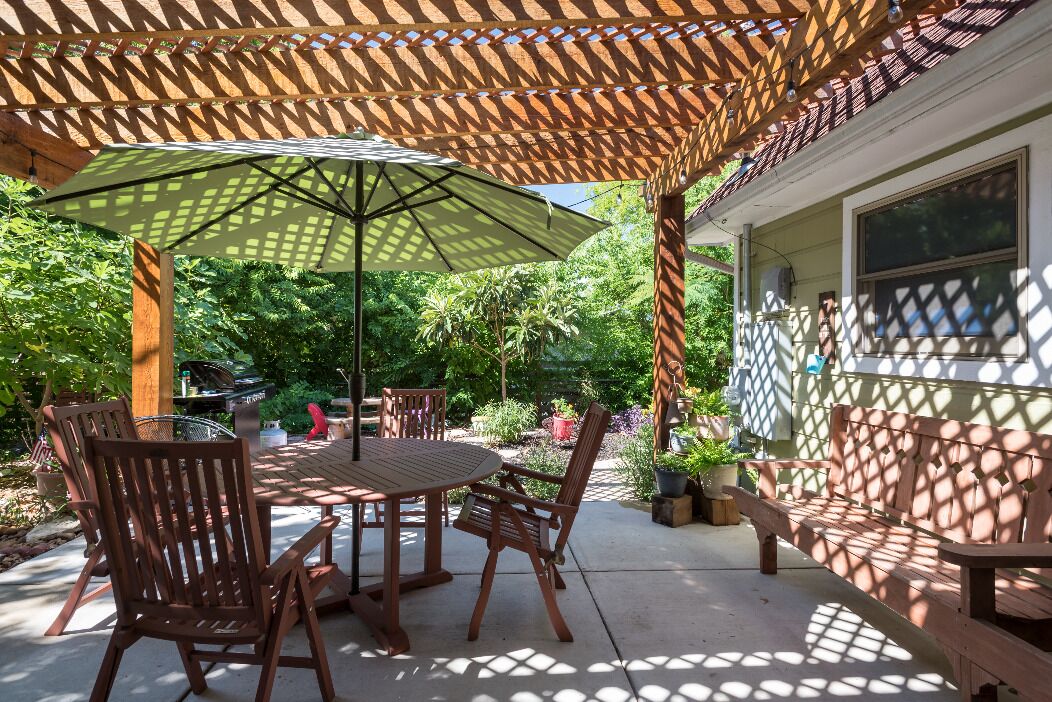 An outdoor patio with a wooden table and chairs, a large umbrella, and a wooden pergola.