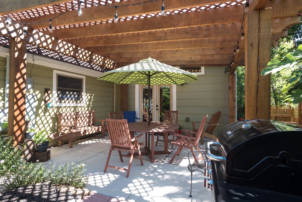 Un patio al aire libre con mobiliario de madera, una parrilla y una pérgola con luces de cadena.