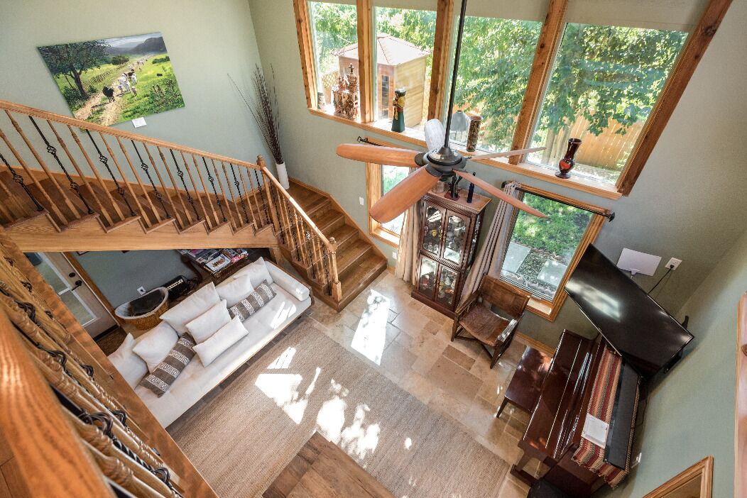 A spacious living area with a wooden staircase, a ceiling fan, a piano, and large windows offering a view of the outdoors.