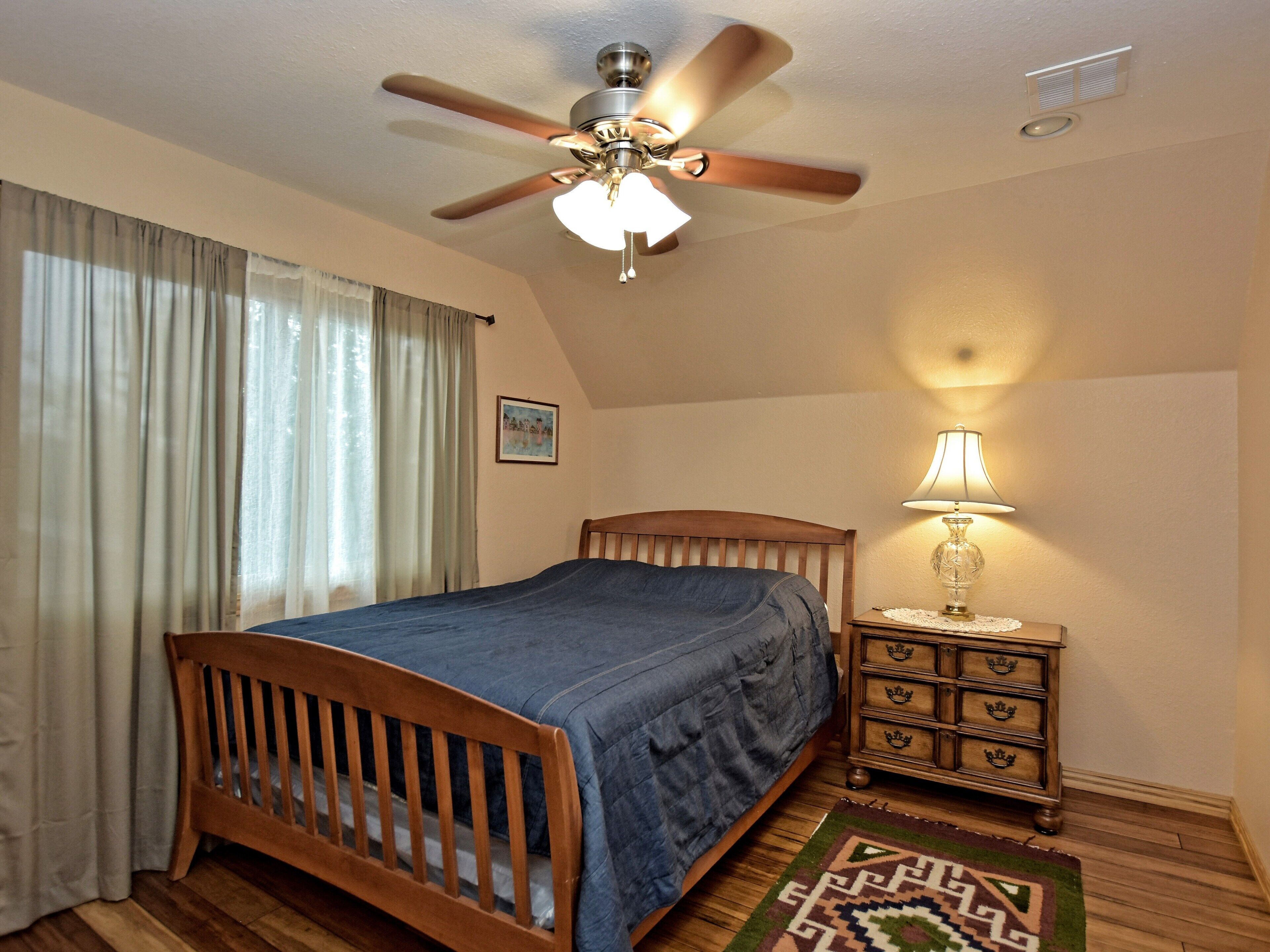 A bedroom with a wooden bed, a ceiling fan, a nightstand with a lamp, and a patterned rug.