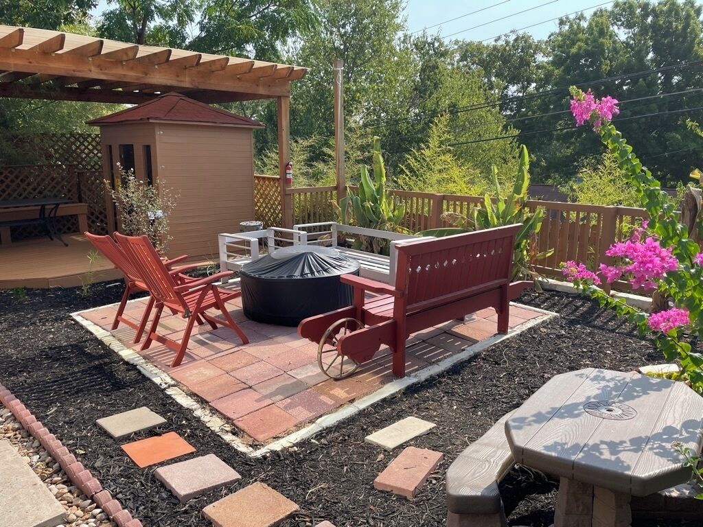 A patio with a wooden pergola, a small shed, red chairs, a fire pit, and a wooden bench.