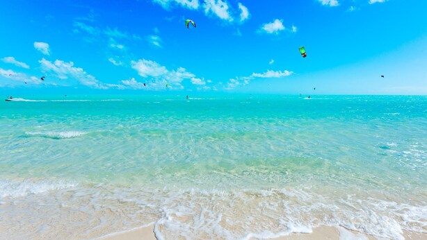 Ein Strand mit klarem türkisblauem Wasser, Sandküste und ein paar Drachen am Himmel.