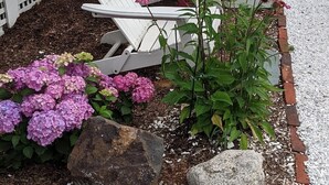 Een rij witte Adirondack-stoelen met een kleine tafel, omringd door roze hortensiakruiden en een bloembed met diverse planten.