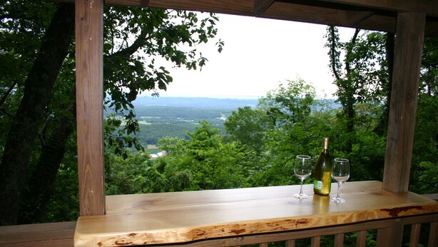 A wooden deck with a bottle of wine and two glasses on a table, overlooking a lush green landscape.