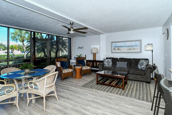 A spacious living area with a large window, wicker furniture, a dark sofa set, and a ceiling fan.