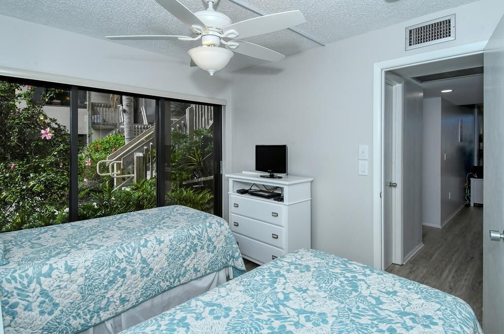 A bedroom with two beds, a ceiling fan, a TV on a white dresser, and a view of a balcony with plants and a staircase.