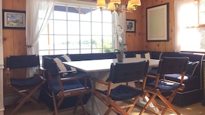 A room with a wooden paneled wall, a large window with curtains, a dining table with chairs, and a chandelier. - Maine Dream Cottage (Biddeford)