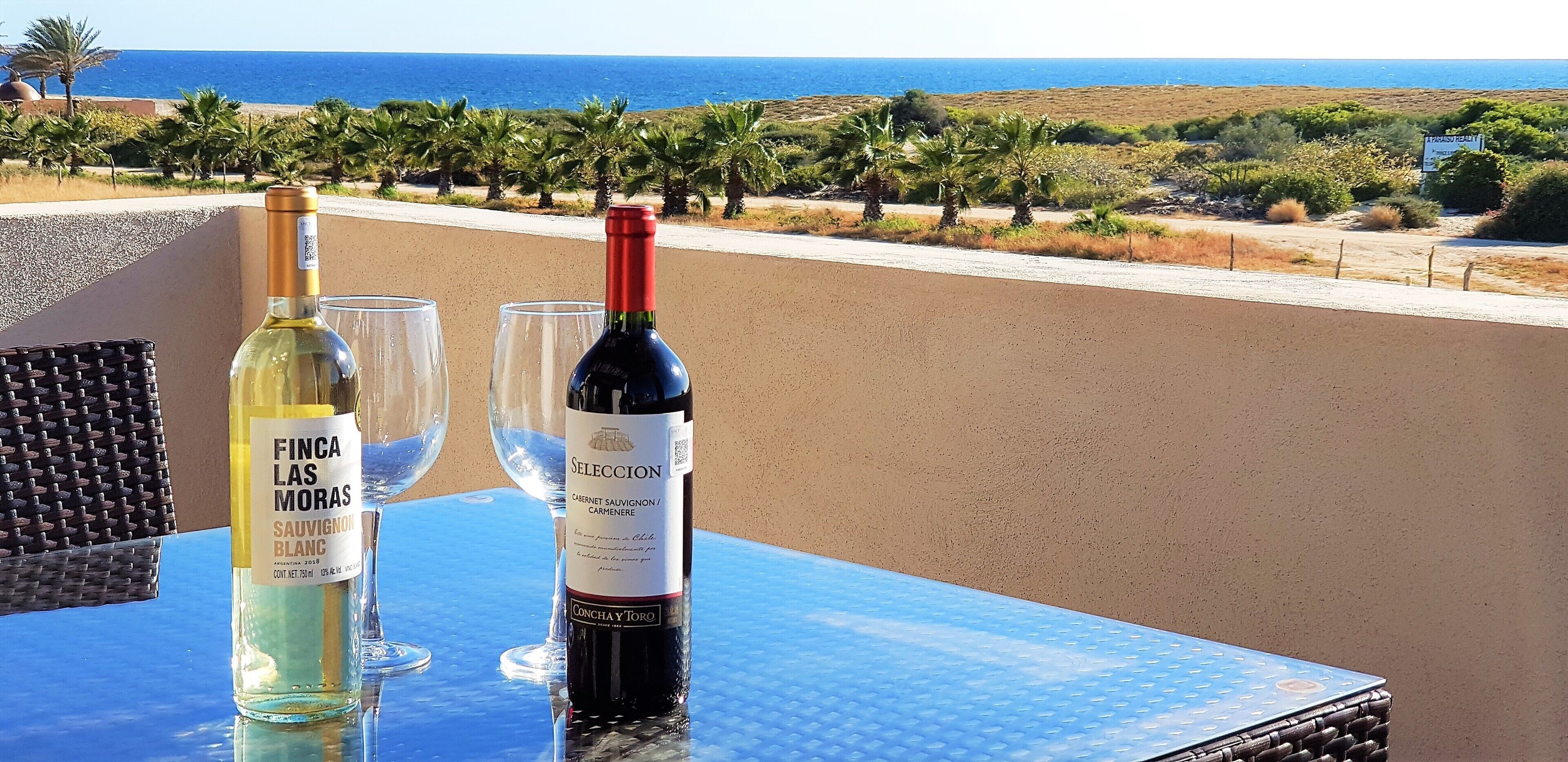 A table with two wine bottles and two glasses on a balcony overlooking a coastal landscape.