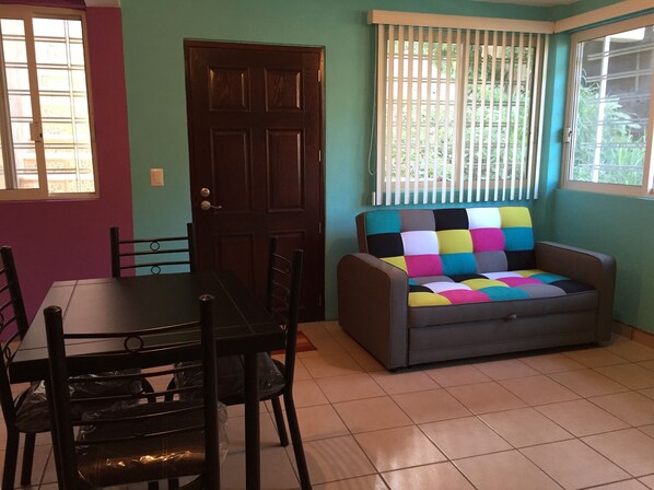 Una habitación con una mesa de comedor y sillas, un sofá con un cojín de colores, una puerta y una ventana con persianas.