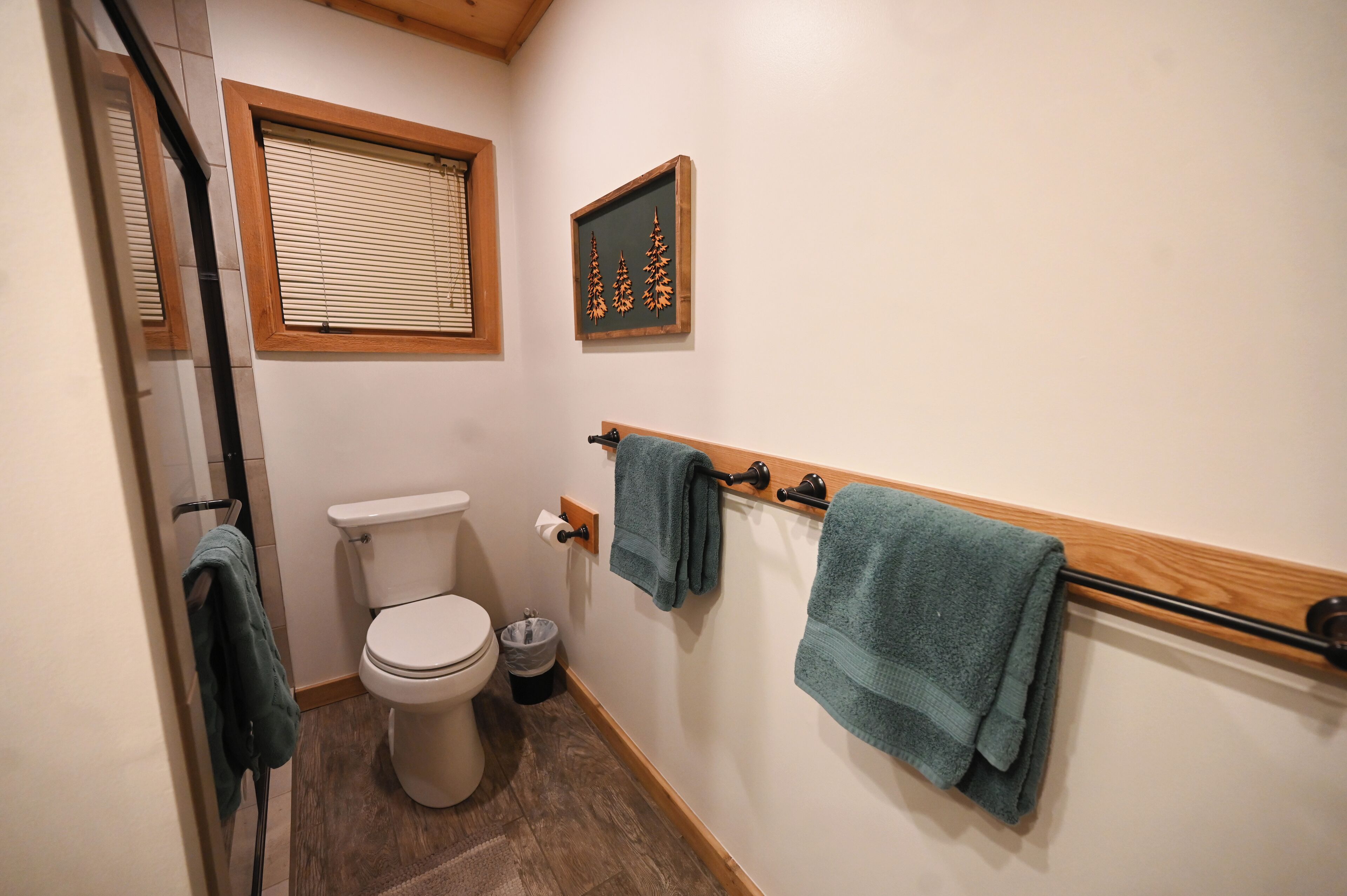 Ein Badezimmer mit Toilette, einem Fenster mit Jalousien, einem gerahmten Bild an der Wand und Handtüchern an einem Halter.