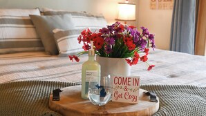 A neatly made bed with striped bedding, a wooden tray with a bottle, a vase of flowers, and a sign that says "COME IN & Get Cozy". - Renovated! Cute, 1 bdrm sleeps 2 Golf/ ski. pool and Wifi! new KING bed. (Mancelona)