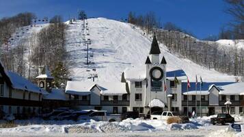 A snow-covered building with a clock tower, ski lift tracks, and a parking area with cars.