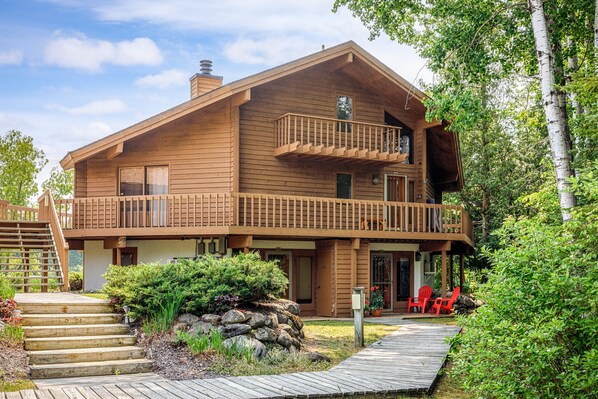 A wooden cabin with a balcony, a stone pathway, and red chairs outside.