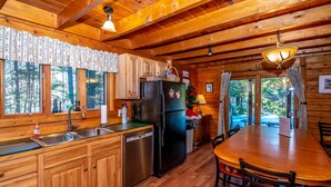 A wooden cabin with a kitchen featuring a refrigerator, sink, and wooden cabinets. There is a dining table with chairs and a view of the forest through large windows.
