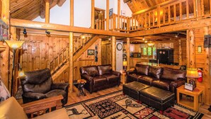 A log cabin interior with a loft bedroom, wooden furniture, and a fireplace.