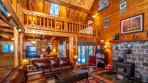 A cozy log cabin interior with a leather sofa, a fireplace, and a wooden staircase.