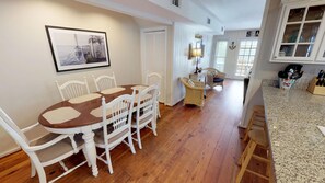 Uma sala de jantar com mesa de madeira e cadeiras brancas, uma cadeira de vime, e uma cozinha com bancadas de granito e vista para o mar.