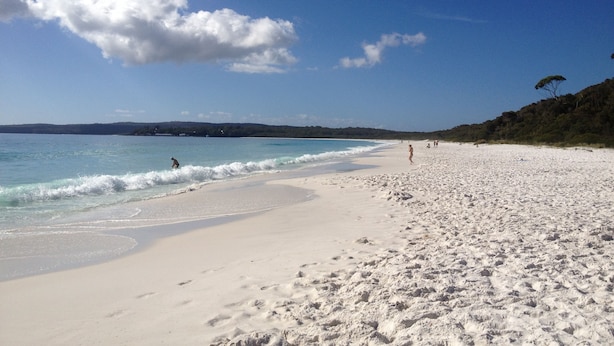 Ein Strand mit weißem Sand, klarem, blauem Wasser und ein paar Personen, die spazieren gehen.