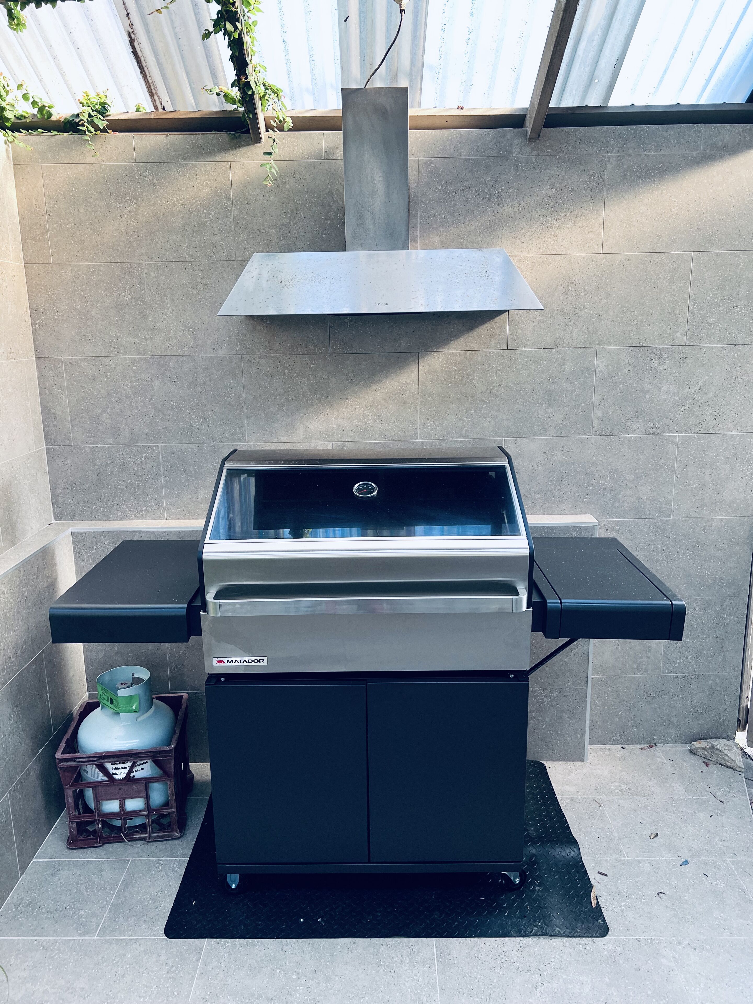 A portable gas grill with a built-in vent hood, a side table, and a gas cylinder storage.