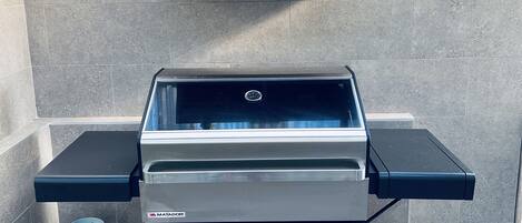 A portable gas grill with a built-in vent hood, a side table, and a gas cylinder storage.