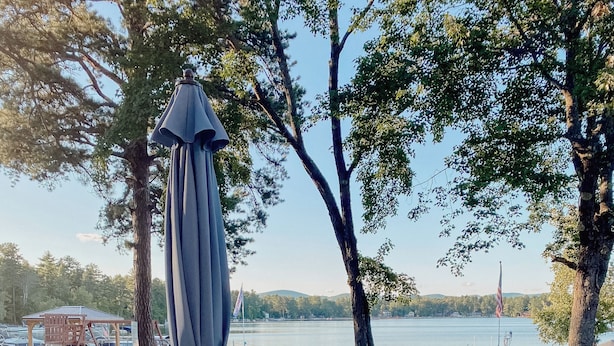 Une terrasse en bois avec une table ronde et des chaises, donnant sur un lac. On aperçoit au loin un belvédère et d’autres chaises.