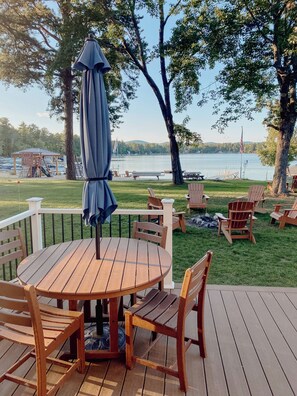 Un terrazzo in legno con un tavolo rotondo e sedie, con vista su un lago e un gazebo con sedie sullo sfondo.