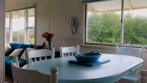 A dining area with a round table, white chairs, a blue tablecloth, and a window with a view of greenery.